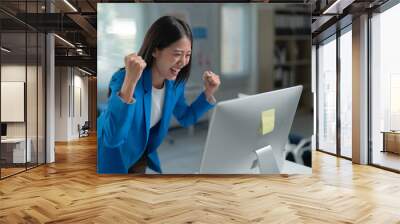 Young businesswoman is raising her fists in the air and smiling while looking at her computer screen in her bright office Wall mural