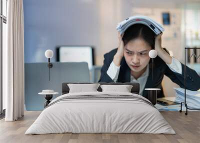 Young businesswoman is suffering from a headache and holding financial documents over her head while working at her desk in the office Wall mural