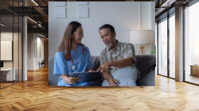 Young female doctor is showing medical information on a tablet to a senior male patient while sitting on a sofa in the living room Wall mural