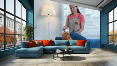 Young housekeeper in an apron cleans a blue sofa in a bright living room with a feather duster, creating a fresh and inviting space Wall mural