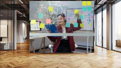 Young manager is using colorful sticky notes to brainstorm ideas and organize her tasks on a transparent glass wall in the office. She is smiling and holding a blue folder in her left hand Wall mural