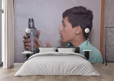 Asian teenage boy drinks water from plastic bottle, wearing a light green shirt with black dots, at home Wall mural