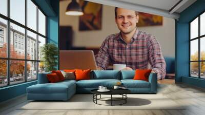 Smiling man in checkered shirt at work, holding mug. Wall mural