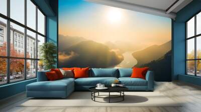 A serene sunrise scene featuring a large sun illuminating a winding river, with a small boat gently floating, surrounded by a landscape of foggy mountains creating a peaceful atmosphere Wall mural