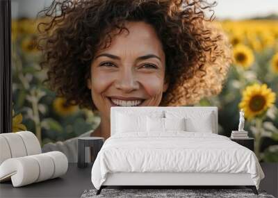 Close-up selfie of a middle-aged mixed-race woman with a bright smile, curly hair, posing in a sunflower field during daytime. Wall mural