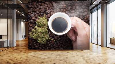 Cannabis coffee. Hand grabbing an ear cup of hot espresso as beside cannabis buds lay on many roasted coffee beans. Wall mural