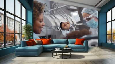 African little girl sitting in dental chair and looking in mirror visiting dentist with mother Wall mural