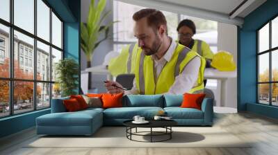 Portrait of male engineer in reflective vest eating lunch and working on laptop in office Wall mural