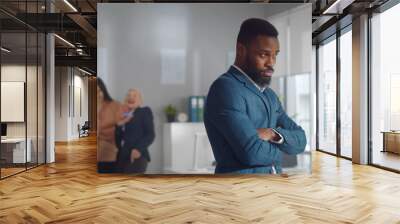 Portrait of sad african businessman standing in office with female colleagues laughing at him on background Wall mural