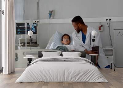 Young african doctor sitting on hospital bed and talking to preteen boy patient Wall mural