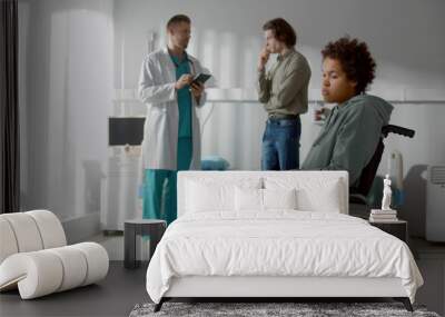 Young upset african woman sitting in wheelchair while doctor consulting her boyfriend on background Wall mural