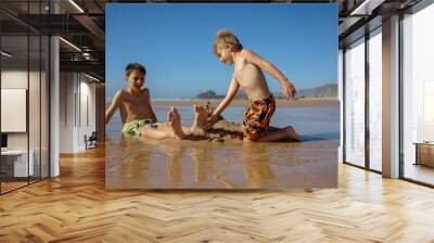 Child, tickling sibling on the beach on the feet with feather, kid cover in sand, smiling, laughing Wall mural