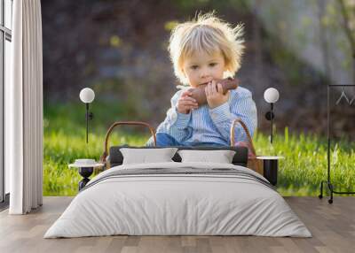 Little toddler boy, eating chocolate bunny in garden on sunset, easter eggs around him Wall mural