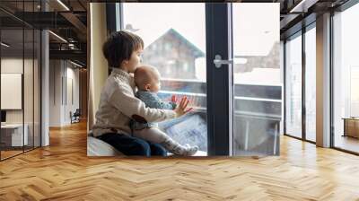 Preschool boy, holding his baby brother, sitting by the window in living room, looking at a snowy landscape outdoors Wall mural