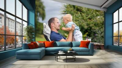 Young father, holding his cute baby boy outdoor, cuddle him, kissing and huging, sitting outdoor on the porch Wall mural