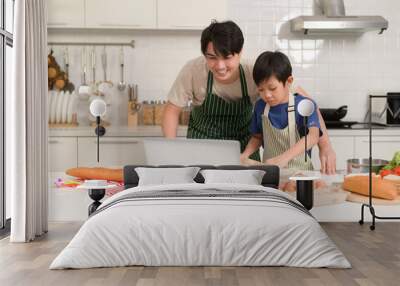 Happy young Asian father and kid are using a laptop computer to learn how to cooking food in their home kitchen. Wall mural