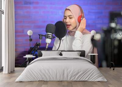 Young smiling muslim female singer wearing headphones with a microphone while recording song in a music studio with colorful lights. Wall mural