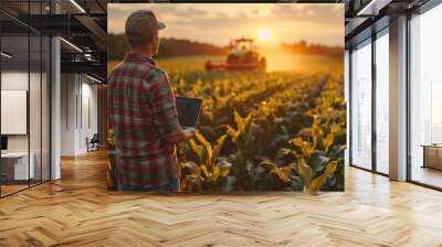 Asian young farmer with laptop standing in corn field, tractor and combine harvester working in corn field in background. Wall mural