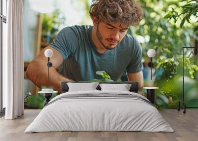 panoramic view of a young, Caucasian man at home using a tiny rake to scratch the ground while sitting next to a potted plant. Wall mural