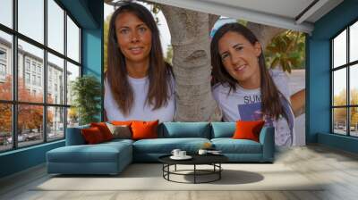 Dos chicas jóvenes que son pareja celebran su aniversario de boda con la bandera LGBTI de fondo en un parque verde y frondoso Wall mural