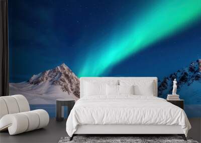 Spectacular view of aurora borealis over snowy mountain range under a starlit night sky, casting a vibrant green glow across the rugged landscape. Wall mural