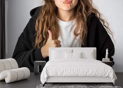 Teenage girl expressing emotions with a thumbs up gesture in casual attire against a neutral background Wall mural