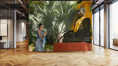 Pretty Asian female in National Thai costume standing pose and pay homage in front of the black-painted Buddha statue outdoors. Wall mural