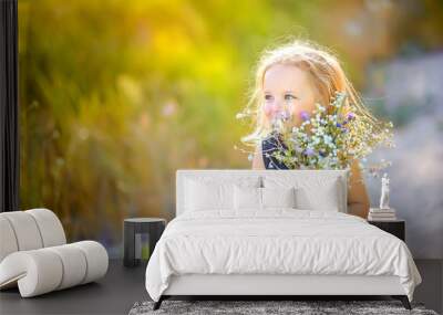 lovely cute little girl with blond hair walks outdoors at sunset, holds a bouquet of wildflowers in her hands, happy childhood Wall mural