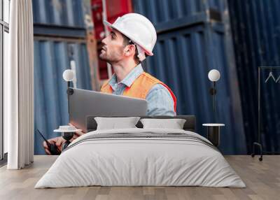 Young confident Caucasian man engineer using laptop computer and wearing white safety helmet and check for control loading containers box from Cargo freight ship for import and export, transport Wall mural