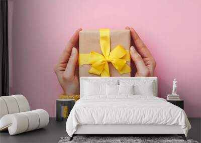 Female hands presenting gift box wrapped in craft paper with soft yellow ribbon bow on pink background, perfect for mother's day, valentine's day, birthday greeting cards, and special occasions with c Wall mural