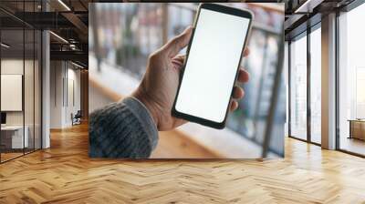 A hand holding a smartphone with a blank screen on a balcony with a city view in the background Wall mural