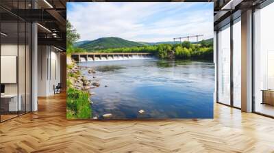 A serene view of a dam, flowing water, and lush greenery under a bright blue sky, surrounded by rolling hills. Wall mural