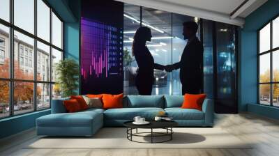 A silhouette of two professionals shaking hands in a modern office, illuminated by colorful digital graphs on a screen. Wall mural