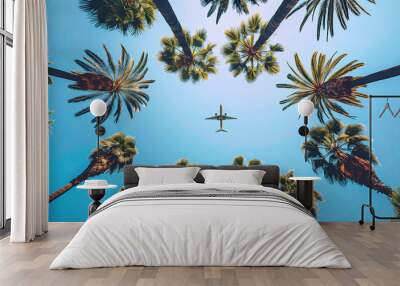 Upward-facing view from ground captures circle of tall palm trees framing clear blue sky with aircraft plane flying overhead. Perspective from below gives dramatic, expansive feel and vacation mood. Wall mural