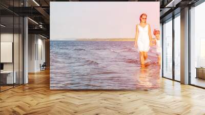 A young mother walks with her daughter on a sandy beach on a summer sunny day at sunset. Wall mural