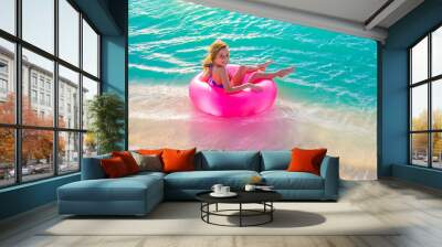 Young blonde girl sitting on a pink lifesaver at the shoreline at the beach Wall mural