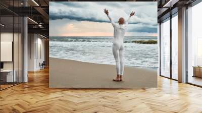 Hairless girl with alopecia in white futuristic suit standing on sea beach stretched out arms to cloudy sky, full length back view portrait, metaphoric performance with bald sensitive teenage girl Wall mural