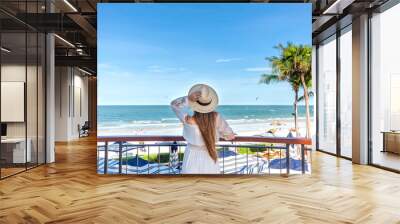 Woman in white dress with straw hat, observing sea view from hotel balcony with palms and beach view. Resort leisure and relaxation. Wall mural