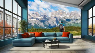 Young man with a hat and a backpack hiking on a trail through green meadows in the Italian Alps. Dolomiti di Brenta mountain peaks are visible in the background. Andalo - Trentino, Italy Wall mural