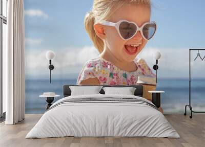 Adorable toddler girl eating ice cream. Portrait of child wearing sunglasses and holding an ice cream with beautiful blue sea and sky behind. Happy summer vacation on the beach. Wall mural