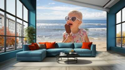 Adorable toddler girl eating ice cream. Portrait of child wearing sunglasses and holding an ice cream with beautiful blue sea and sky behind. Happy summer vacation on the beach. Wall mural