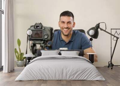 Young smiling cheerful man sitting at the office desk in front of the camera making a video. Male blogger recording a video tutorial. Wall mural