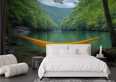 A yellow hammock slung between two trees overlooking a calm, green lake nestled within a verdant valley. Wall mural