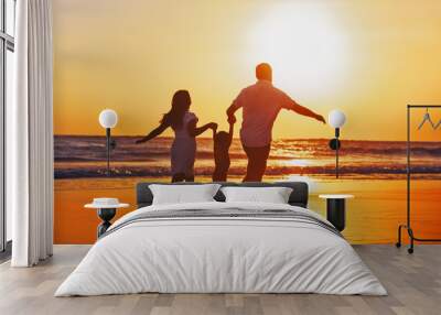 Happy family - father, mother, baby son hold hands and run with fun along edge of sunset sea on black sand beach. Active parents and people outdoor activity on tropical summer vacations with children. Wall mural