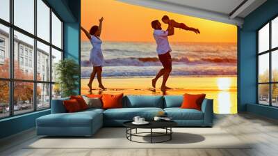 Happy family - father, mother, baby son walk with fun along edge of sunset sea surf on black sand beach. Active parents and people outdoor activity on summer vacations with children on Bali island Wall mural