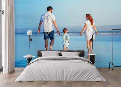 Happy family - young father, mother, baby son strolling together, child run with fun by water pool along sunset sea surf on tropical beach. Travel lifestyle, people walking with kid on summer vacation Wall mural