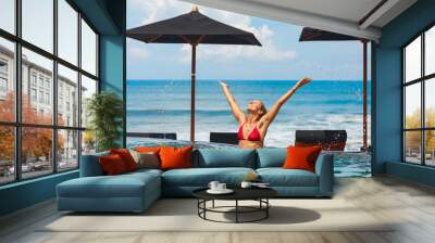 Happy woman have fun in outdoor infinity pool with view to ocean surf. Positive girl swim, relax at poolside, spread hands high with water splashes. Healthy lifestyle, people on summer beach holiday Wall mural