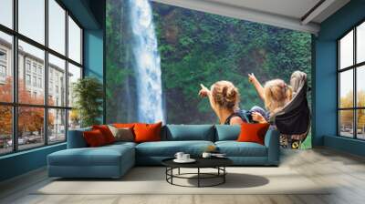 In waterfall pool young happy mother hold little traveller - baby girl in carrying backpack on back. Travel adventure, hiking activity with child. Lifestyle on family summer vacation and weekend tour Wall mural