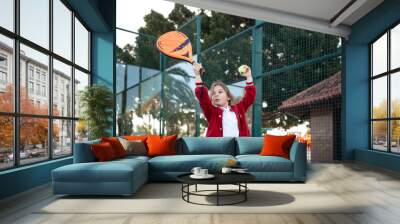 An action shot of a child reaching high to serve a padel tennis ball on an outdoor court, with palm trees in the background Wall mural