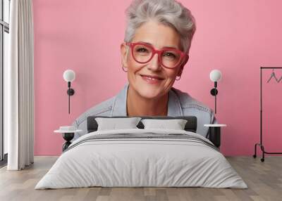 A woman wearing pink glasses and a blue jacket is smiling. She looks happy and content. The pink background adds a pop of color to the scene, making it feel cheerful and uplifting Wall mural